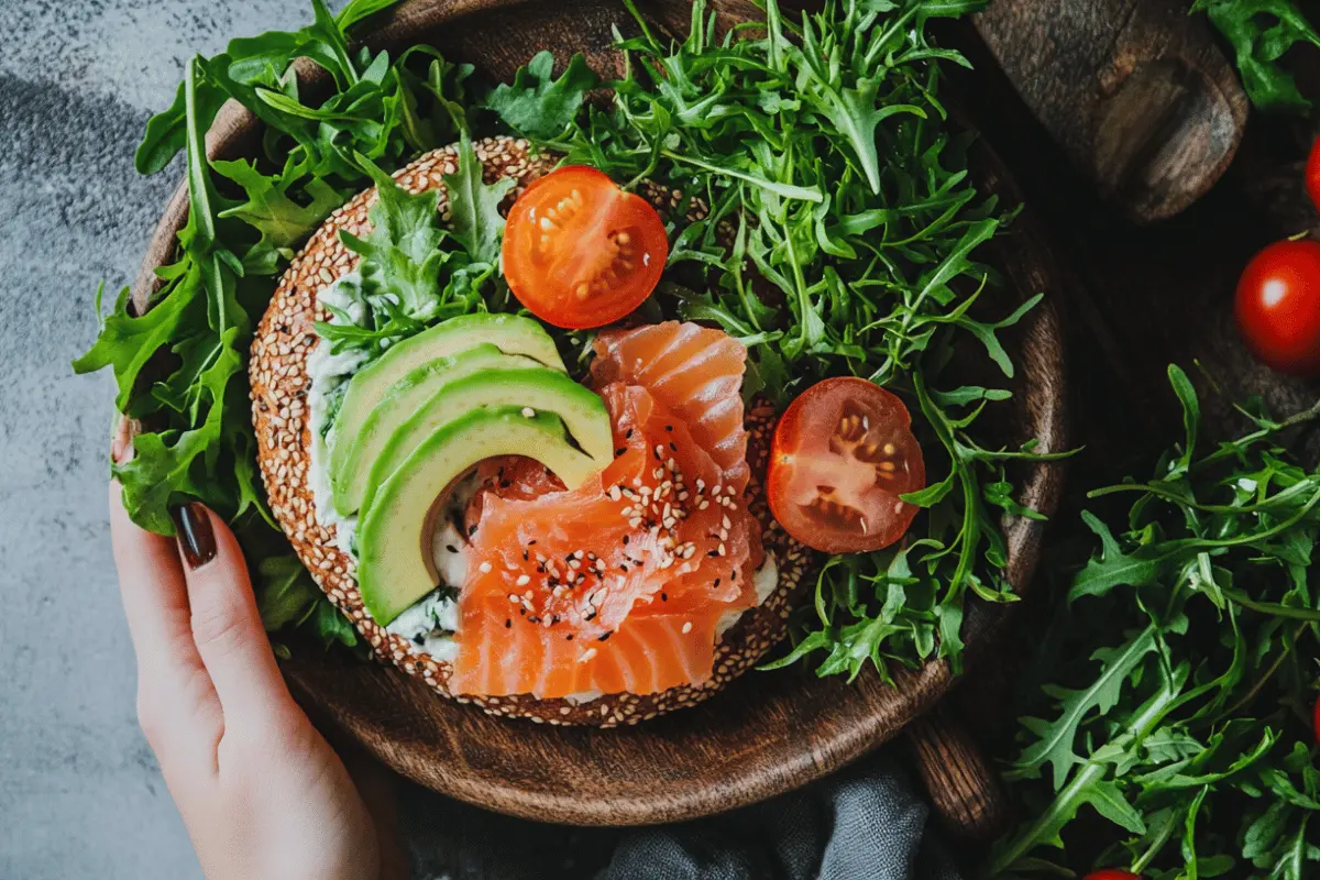 A delicious bagel with cream cheese, topped with fresh avocado slices, smoked salmon, cherry tomatoes, and sesame seeds, served on a wooden plate with a side of arugula. Is A Bagel With Cream Cheese Healthy? Find out more!